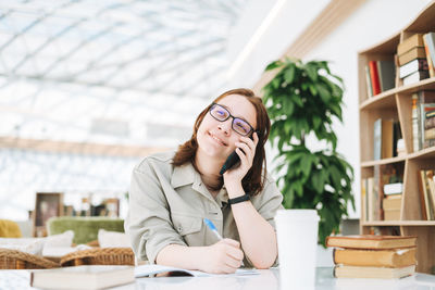 Young brunette teenager girl student in glasses using mobile phone working at green modern library 