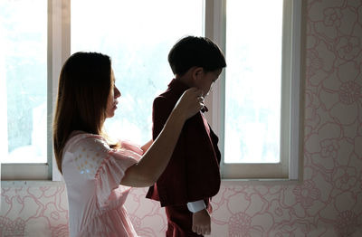 Side view of woman dressing up son by window at home
