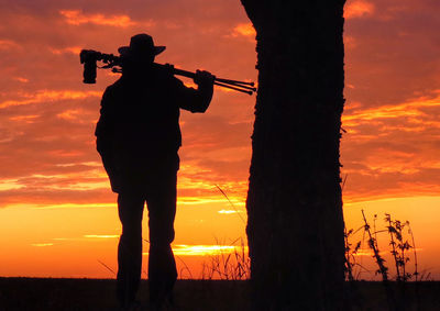 Rear view of silhouette man carrying tripod against orange sky during sunset