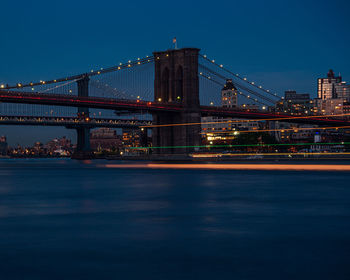 View of suspension bridge in city at night with a ship passing by 