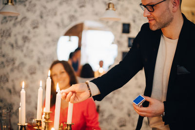 Smiling man igniting candles at dinner party with friend in background