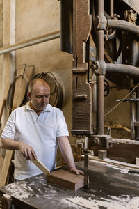 Talented male woodworker using band saw for cutting wooden plank in grungy workshop