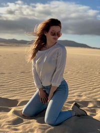 Full length of young woman kneeling on sand at desert against sky