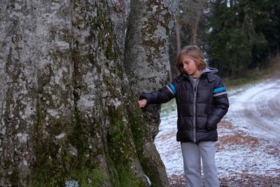 Boy standing by tree trunk on roadside during winter