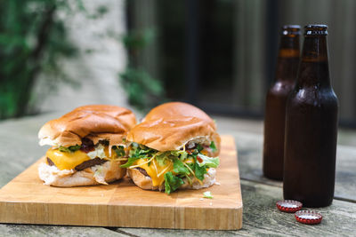 A set of cheeseburgers on a wooden plate and a couple of beers on an outdoor table. suburban.