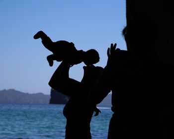 Silhouette mother kissing child while standing by sea against clear blue sky