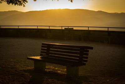 Empty bench by railing against sky during sunset