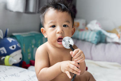 Portrait of cute baby boy holding stethoscope while sitting on bed at home