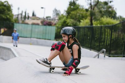 Side view of playful girl sitting on skateboard at sports ramp