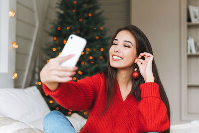 Young asian woman in red sweater taking selfie by mobile on bed in room with christmas tree at home