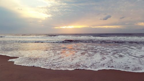 Scenic view of waves rushing at sea shore against cloudy sky during sunset