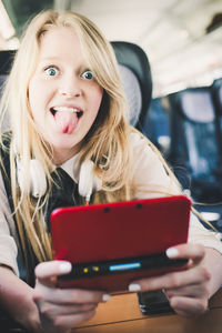 Portrait of cheerful teenage girl sticking out tongue while using mobile phone in train