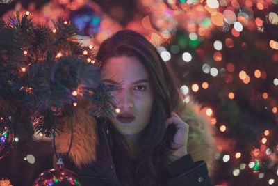 Portrait of young woman standing against illuminated christmas tree at night