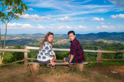 Portrait of couple sitting on bench at mountain peak against sky