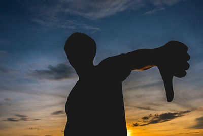 Silhouette man gesturing thumbs down against sky during sunset