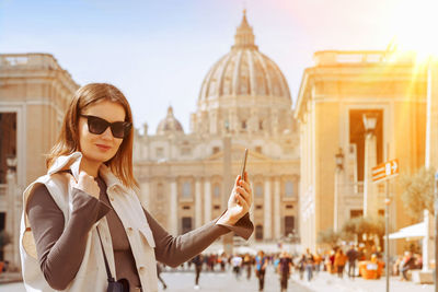 Cheerful beautiful young woman takes a selfie on a smartphone while walking in rome 