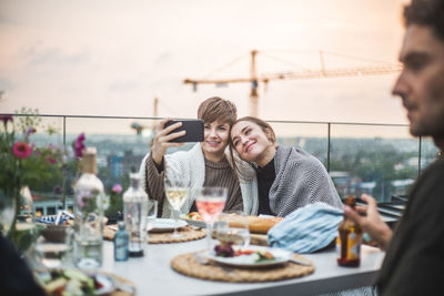 Smiling female friends taking selfie with blanket while sitting at rooftop during social gathering