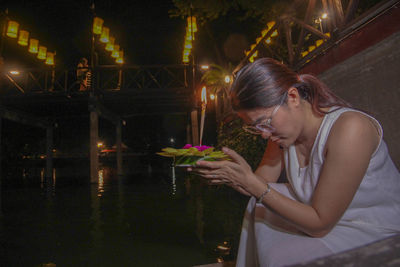 Side view of woman looking down while sitting on illuminated lamp at night