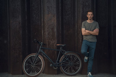 Man leaning aginst metal wall with arms crossed by bicycle
