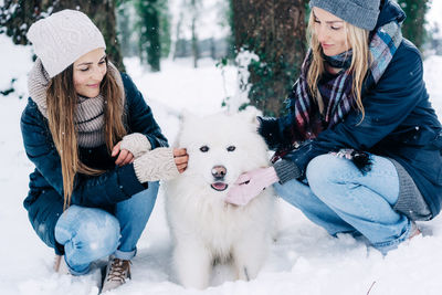 Two female friends walking in the park in the snow play with a dog.