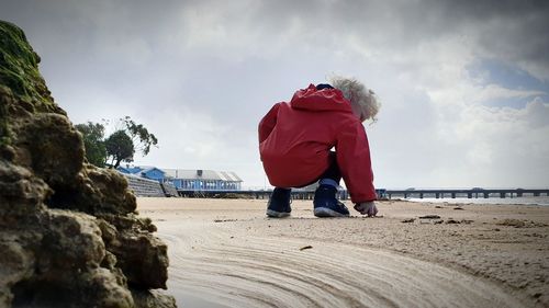 Rear view of boy crouching at beach against sky