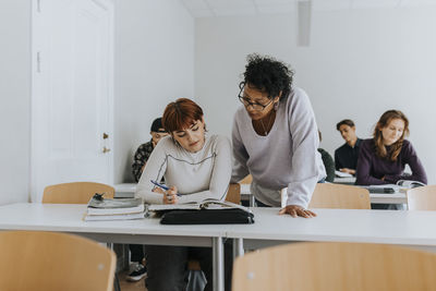 Mature female teacher assisting teenage student at desk in classroom