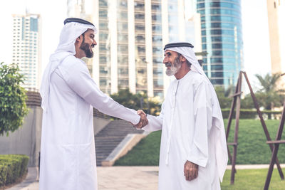 Man holding hands of grandfather wearing dish dash on footpath at park in city