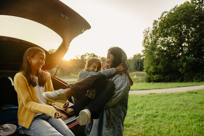 Cheerful woman carrying daughter near electric car trunk on sunset