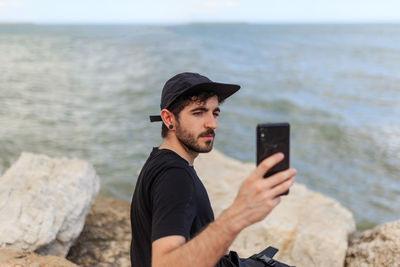 From above side view of bearded male in cap and black clothes taking photo with cellphone while sitting on rough stones near ocean