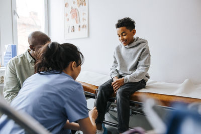Female pediatrician checking knee of boy with reflex hammer in medical examination room
