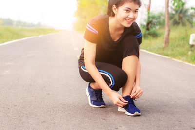 Smiling young woman tying shoelace while crouching on road