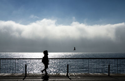 Silhouette woman waking on footpath by sea against cloudy sky