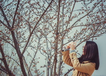 Woman taking selfie through mobile phone against bare tree