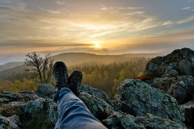 Low section of man on rock against sky during sunset