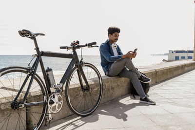 Man using mobile phone while sitting on retaining wall by bicycle during sunny day