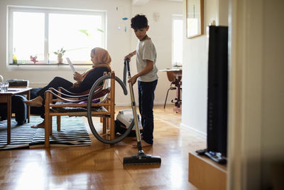 Boy cleaning hardwood floor with vacuum cleaner while mother sitting on armchair at home