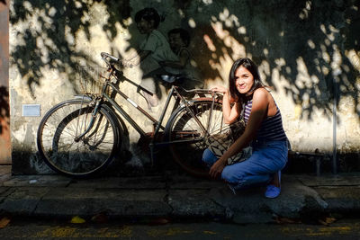 Portrait of young woman crouching by bicycle against wall