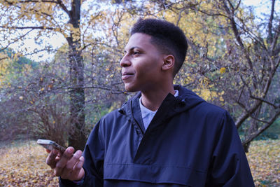 Young man looking away while holding mobile phone against tree