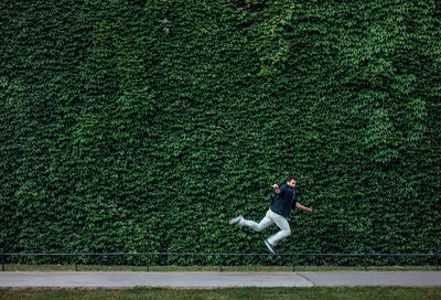 Side view of young man jumping on footpath by ivy wall