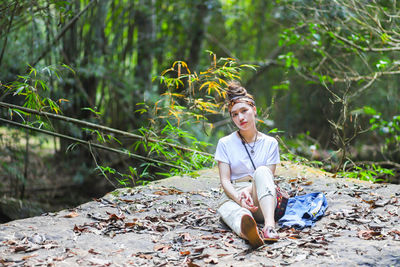 Portrait of serious young woman sitting on rock against trees in forest