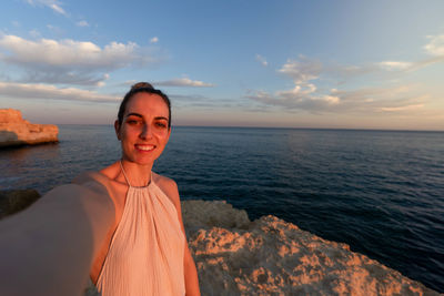 Portrait of young woman standing at beach against sky during sunset