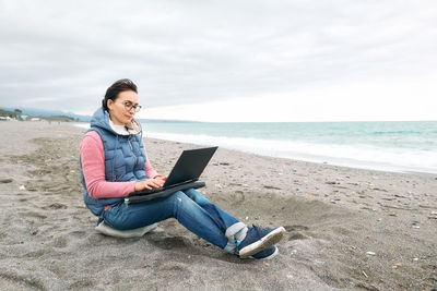 Beautiful sporty woman with laptop computer working outdoors while sitting on winter beach. 