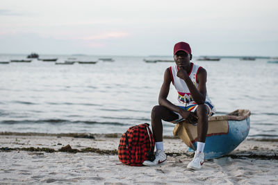 Portrait of young man sitting on shore at beach against sky