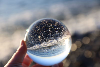 Cropped hand holding crystal ball against sea during sunny day