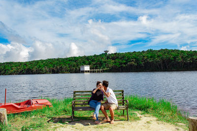 Woman sitting on bench by lake against sky