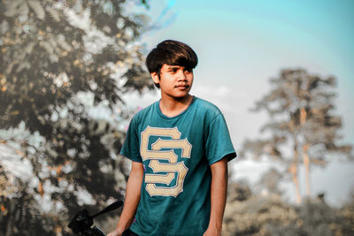 Young man looking away while standing against trees