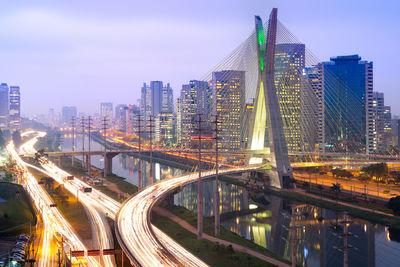 Illuminated bridge by buildings against sky in city