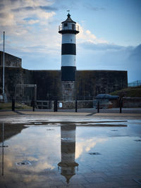 Reflection of lighthouse in puddle on building against sky