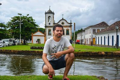 Portrait of young man crouching by canal against building