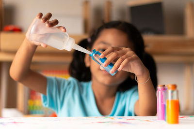 Cute girl pouring water in color bottle on table at home